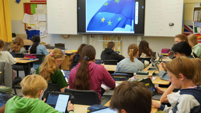 Schüler:innen arbeiten an ihren Ipads. Vor ihnen ist auf einem Whiteboard eine Europaflagge zu erkennen.
