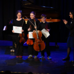 Drei Musiker stehen auf einer Bühne und halten Urkunden in den Händen
