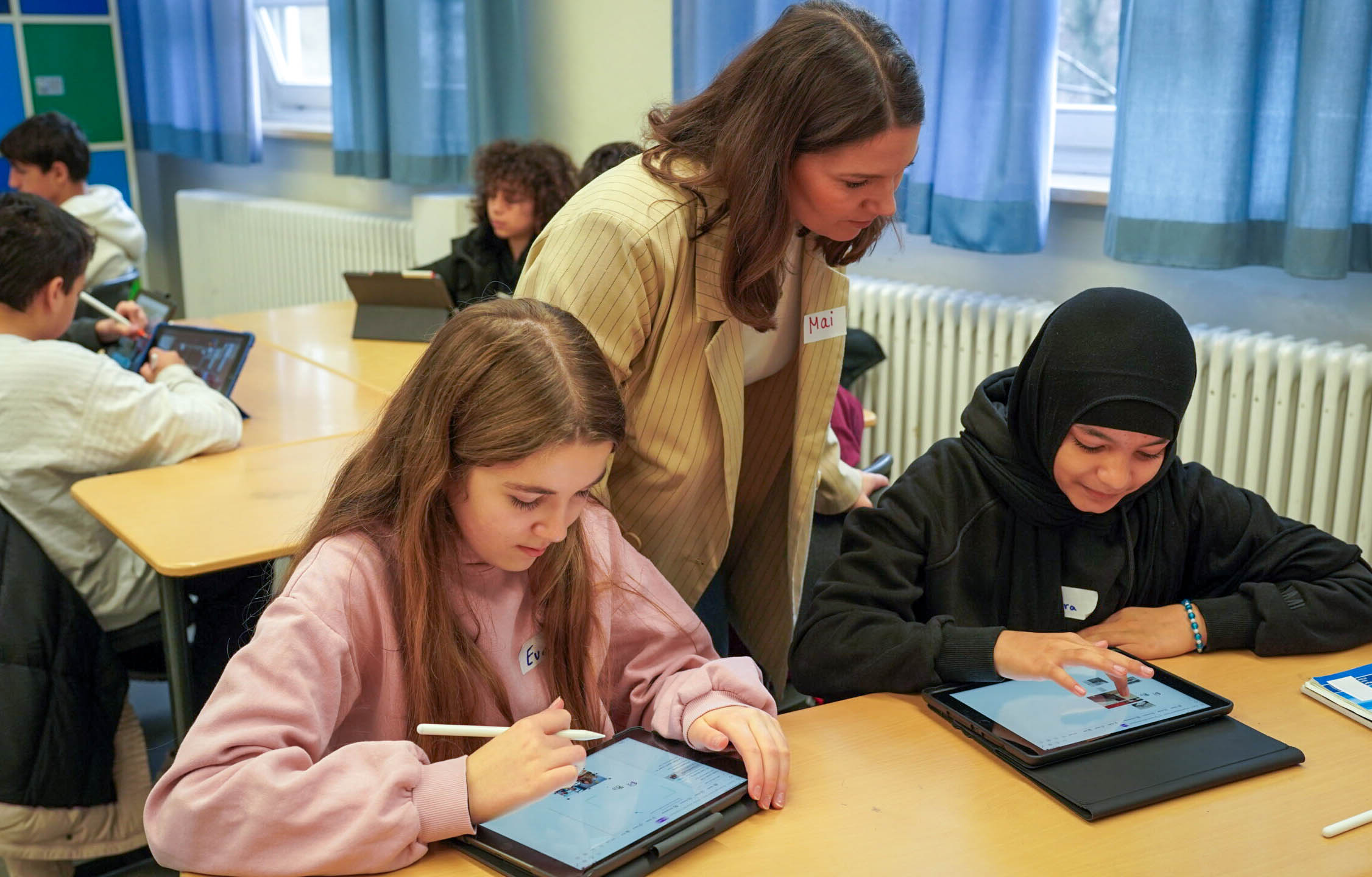 Schüler:innen arbeiten an Aufgaben, Lehrerin hilft dabei